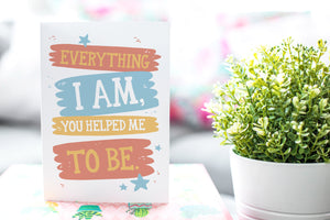 A photo of a card featured on a tabletop next to a white planter filled with a green plant. ​​The card features the words “Everything I Am, You Helped Me To Be.”