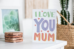 A greeting card featured standing up on a white tabletop with a framed photo of a succulent in the background and a stack of wooden coasters. There’s a woven basket in the background with a cactus inside. The card features illustrated lettering reading “I love you Mum” with small hearts around the words.