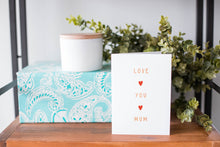 Load image into Gallery viewer, A greeting card is on a table top with a present in blue wrapping paper in the background. On top of the present is a candle and some greenery from a plant too. The card features the words “Love You Mum” with a small, red heart in between each word. 