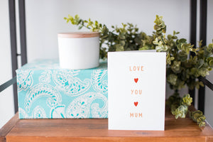 A greeting card is on a table top with a present in blue wrapping paper in the background. On top of the present is a candle and some greenery from a plant too. The card features the words “Love You Mum” with a small, red heart in between each word. 