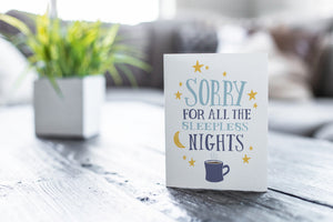 A greeting card featured on a black, wood coffee table. There’s a white planter in the background with a green plant. There’s also a gray sofa in the background with a white pillow. The card features the words “Sorry for all the sleepless nights.”