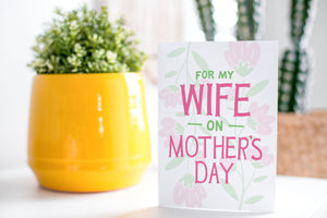 A greeting card is on a table top with a yellow plant pot and a green plant inside. The card features illustrated flowers in the background with the words “For my wife on Mother’s Day.”