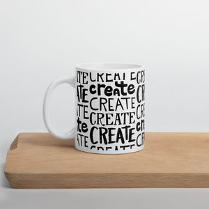 A white mug sitting on a piece of wood. The quote on the mug says “create" repeated over and over in different hand lettered fonts. 