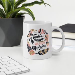 A mug featured on a desk with a plant and a keyboard. The white mug features hand drawn lettering with the words "Tender Warriors Are Being Made Here" The words are in pink, navy and dark mustard yellow. 