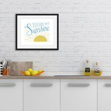 Load image into Gallery viewer, A white print in a black frame hangs on a kitchen wall. The wall is white painted brick with a wooden worktop holding utensils, chopping boards, bottles of oil, and lemons. The print reads &#39;You are my sunshine&#39; in blue lettering, over a yellow sun illustration