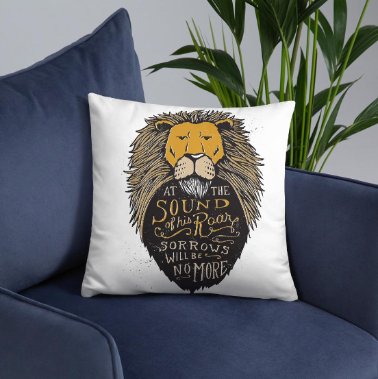 A white square pillow sitting on a blue sofa chair with a plant in the background. The pillow artwork features hand drawn illustration of the Chronicles of Narnia lion character Aslan. Inside the illustration there is the quote “At The Sound of Your Roar, Sorrows Will Be No More.”