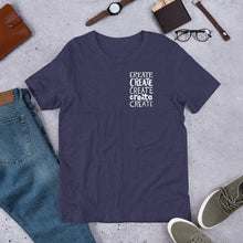 Load image into Gallery viewer, A short sleeved T-shirt laying flat with objects around it. The tee is in the color heather midnight navy blue with the words &quot;create, create, create, create, create&quot; in white in a small rectangle on the upper left side. 