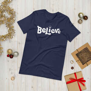 A navy T-shirt laying on the ground with Christmas items surrounding it. The T-shirt features the word Believe in the middle in white lettering with the "I" of the word featured as a illustrated Christmas tree. 