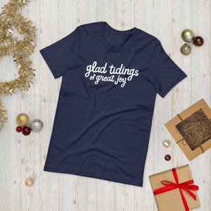 A navy blue T-shirt laying on the ground with Christmas items surrounding it. The T-shirt features the words  "glad tidings of great joy" in white. 