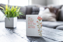 Load image into Gallery viewer, A greeting card featured on a black, wood coffee table. There’s a white planter in the background with a green plant. There’s also a gray sofa in the background with a white pillow. The card features illustrated flowers on the right hand side of the card. The petals are muted pink and the stem and leaves are green and light brown. To the left of the flowers the card reads &quot;I have fallen more in love with you since you became a mother.&quot; 