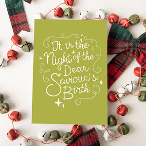 A Christmas card featured on top of some red and white Christmas decorations. The background of the card is a lime green with the word "it is the night of the dear saviour's birth" in script white lettering.