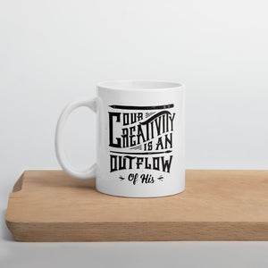 A white mug sitting on a piece of wood. The white mug features hand drawn lettering with the words "Our creativity is an outflow of His." The words are in black.
