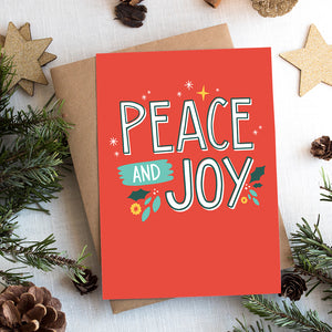 A photo of a Christmas card on top of a brown paper wrapped gift with Christmas decor around it. The card has a red background with the words "peace and joy" in white and illustrations of stars and holly leaves around the wording.