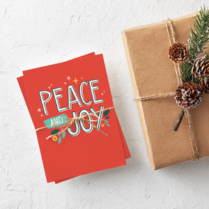 A stack of Christmas cards with brown string wrapped around them. A brown craft paper gift is off to the side. The card has a red background with the words "peace and joy" in white and illustrations of stars and holly leaves around the wording.