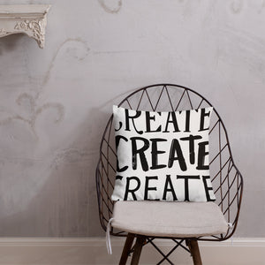 A pillow on a chair against a grey wall. The pillow is white and features the words “create, create, create" in black lettering with each word in three different fonts. 