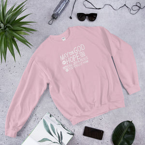 A light pink sweatshirt laying on the ground with objects around it. The sweatshirt features hand drawn lettering in white with the words "May the God of hope fill you with all joy and peace as you trust him."