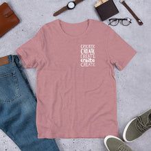 Load image into Gallery viewer, A heather orchid pinnk T-shirt laying flat with objects around it. The tee features the lettering and illustration in white. The phrase &quot;create, create, create, create, create&quot; is in a small rectangle on the upper left side. 