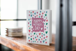 A card on a wood tabletop with an object in the background that is out of focus. The card features the words “Happy Mother’s Day.”