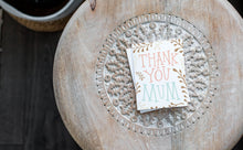 Load image into Gallery viewer, A greeting card laying on a wooden table with some cut wood details. The card features illustrated plant leaves around the words “Thank you Mum.”