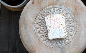 A greeting card laying on a wooden table with some cut wood details. The card features illustrated plant leaves around the words “Thank you Mum.”