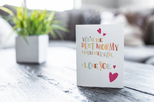 A greeting card featured on a black, wood coffee table. There’s a white planter in the background with a green plant. There’s also a gray sofa in the background with a white pillow. The card features the words “You’re the best mommy I could ever have. I love you.” 