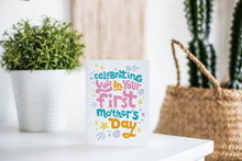 Load image into Gallery viewer, A greeting card is featured on a white tabletop with a white planter in the background with a green plant. There’s a woven basket in the background with a cactus inside. The card features the words “Celebrating you on your first Mother’s Day.”