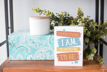 Load image into Gallery viewer, A greeting card is on a table top with a present in blue wrapping paper in the background. On top of the present is a candle and some greenery from a plant too. The card features the words “Everything I Am, You Helped Me To Be.”