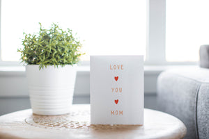 A greeting card laying on a wooden table with some cut wood details. The card features the words “Love You Mom.”
