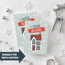 Load image into Gallery viewer, Two Christmas cards laying on a white background with white and silver Christmas decorations on the table. The Christmas card has a light blue background with stars. The words &quot;this house believes&quot; is featured above an illustrated house. The words and house are in white, green and light blue. In the lefthand bottom corner is a navy arrow with the words &quot;download &amp; print, send to loved ones&quot; over the image.