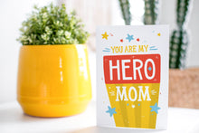 Load image into Gallery viewer, A greeting card is on a table top with a yellow plant pot and a green plant inside. The card features the words “You are my hero mom.”