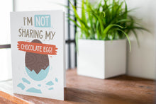 Load image into Gallery viewer, A greeting card is featured on a desktop with a green plant in the background. The card features an illustrated chocolate Easter egg with the words “I’m not sharing my chocolate egg!”