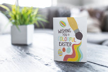 Load image into Gallery viewer, A greeting card featured on a black, wood coffee table. There’s a white planter in the background with a green plant. There’s also a gray sofa in the background with a white pillow. The card features an illustrated paint brush and Easter eggs with the words “Wishing you a colorful Easter.”