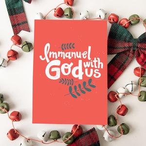 A Christmas card featured on top of some red and white Christmas decorations. The card has a light red background with the words "Immanuel God with Us" in white with a couple of plant leaves in navy around the words. 