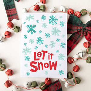 A Christmas card featured on top of some red and white Christmas decorations. The card has a white background with the words "let it snow" at the bottom in red. Illustrated snowflakes are on the rest of the card in different shades of blue.