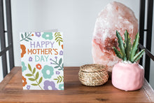 Load image into Gallery viewer, A card on a wood tabletop and on the right side of the card is a woven basket, a pink plant pot with a cactus in it and a pink crystal rock. The card features the words “Happy Mother’s Day” with illustrated flowers around the words. 
