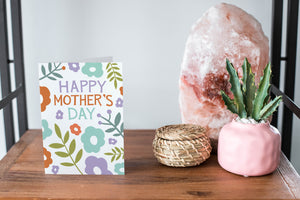 A card on a wood tabletop and on the right side of the card is a woven basket, a pink plant pot with a cactus in it and a pink crystal rock. The card features the words “Happy Mother’s Day” with illustrated flowers around the words. 