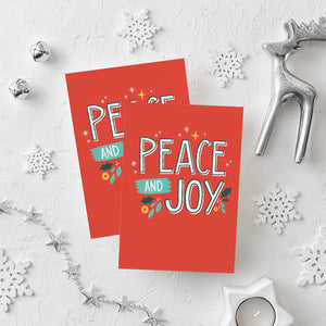 Two Christmas cards laying on a white background with white and silver Christmas decorations on the table. The card has a red background with the words "peace and joy" in white and illustrations of stars and holly leaves around the wording. 