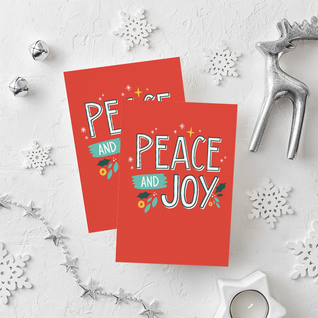 Two Christmas cards laying on a white background with white and silver Christmas decorations on the table. The card has a red background with the words 