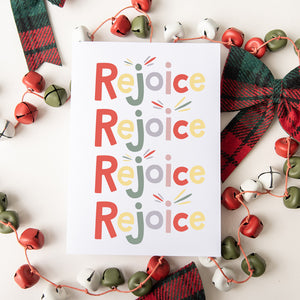 A Christmas card featured on top of some red and white Christmas decorations. The card has a white background and features the word "rejoice" repeated four times. The letters of the word are in different colors of muted red, yellow, green, purple and pink.