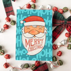 A Christmas card featured on top of some red and white Christmas decorations. The card has a blue background with the words 'ho ho ho' in a lighter shade of blue. On top of the background is an illustrated Santa Claus with the words "Merry Christmas" in his beard.