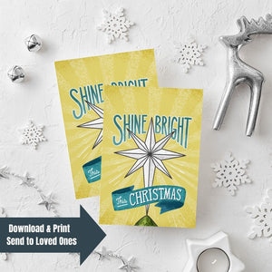 Two Christmas cards laying on a white background with white and silver Christmas decorations on the table. The Christmas card has a yellow background with the words 'shine bright this Christmas' in blue and white. There's an illustrated vintage star Christmas tree topper featured in between the words. In the lefthand lower corner is a navy arrow with the words "download & print, send to loved ones" over the image.