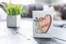 Load image into Gallery viewer, A greeting card featured on a black, wood coffee table. There’s a white planter in the background with a green plant. There’s also a gray sofa in the background with a white pillow. The card features an illustrated Easter bunny holding a heart with the words “some bunny loves you.”