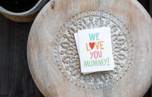 Load image into Gallery viewer, A greeting card laying on a wooden table with some cut wood details. The card features the words “We Love You Mummy!” with the “O” in the word love featured as a heart.