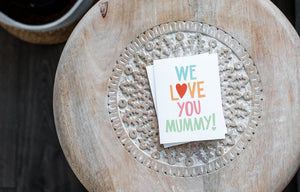 A greeting card laying on a wooden table with some cut wood details. The card features the words “We Love You Mummy!” with the “O” in the word love featured as a heart.