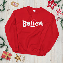 Load image into Gallery viewer, A red sweatshirt laying on a table with Christmas objects around it. The sweatshirt features the word Believe in white with an illustrated Christmas tree as the &quot;I.&quot;