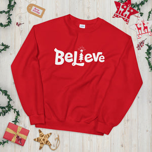 A red sweatshirt laying on a table with Christmas objects around it. The sweatshirt features the word Believe in white with an illustrated Christmas tree as the "I."