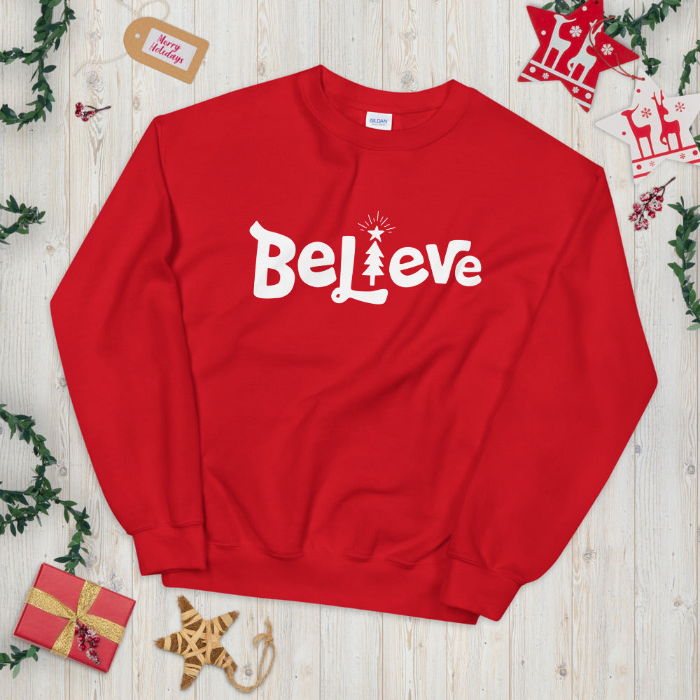 A red sweatshirt laying on a table with Christmas objects around it. The sweatshirt features the word Believe in white with an illustrated Christmas tree as the 