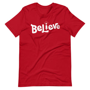 A red T-shirt on a white background. The red shirt features the hand lettered word "Believe" in white with the "I" featured as an illustrated Christmas tree. 