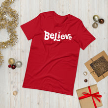 Load image into Gallery viewer, A red T-shirt laying on the ground with Christmas items surrounding it. The T-shirt features the word Believe in the middle in white lettering with the &quot;I&quot; of the word featured as a illustrated Christmas tree. 