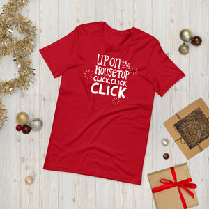 A red T-shirt laying on the ground with Christmas items surrounding it. The T-shirt features the words  "Up on the housetop, click, click, click" in white. There are three stars around the words in white. 